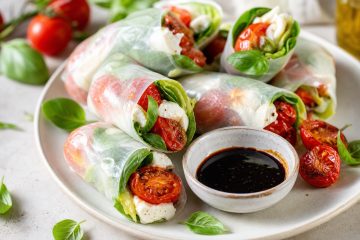 An editorial-style food photograph of Caprese summer rolls made with translucent rice paper, neatly arranged on a light ceramic plate or wooden board, each roll showing fresh basil, crisp romaine, torn mozzarella, and glossy roasted grape tomatoes visible through the wrapper; a small bowl of thick reduced balsamic vinegar for dipping beside them, with a few roasted tomato halves and basil leaves scattered naturally around the composition. Bright natural window light, fresh summer brunch mood, clean minimal styling, soft shadows, shallow depth of field, vibrant red, green, white, and amber tones, high-resolution, appetizing, elegant, no text, no hands, no branding.