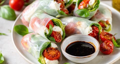 An editorial-style food photograph of Caprese summer rolls made with translucent rice paper, neatly arranged on a light ceramic plate or wooden board, each roll showing fresh basil, crisp romaine, torn mozzarella, and glossy roasted grape tomatoes visible through the wrapper; a small bowl of thick reduced balsamic vinegar for dipping beside them, with a few roasted tomato halves and basil leaves scattered naturally around the composition. Bright natural window light, fresh summer brunch mood, clean minimal styling, soft shadows, shallow depth of field, vibrant red, green, white, and amber tones, high-resolution, appetizing, elegant, no text, no hands, no branding.