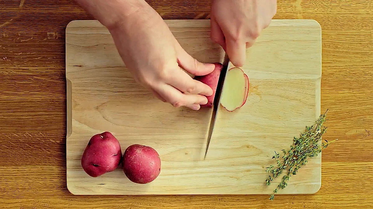 Red potatoes being sliced into thin rounds on a cutting board